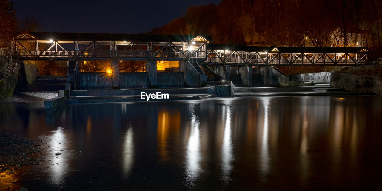 ILLUMINATED BRIDGE OVER RIVER BY BUILDINGS