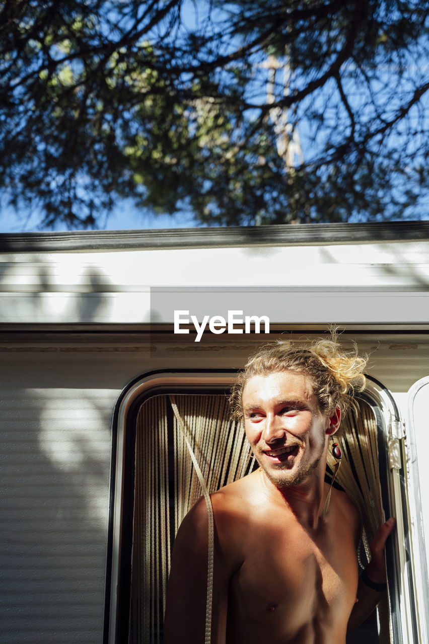 Smiling shirtless man with blond hair standing at doorway of camper van
