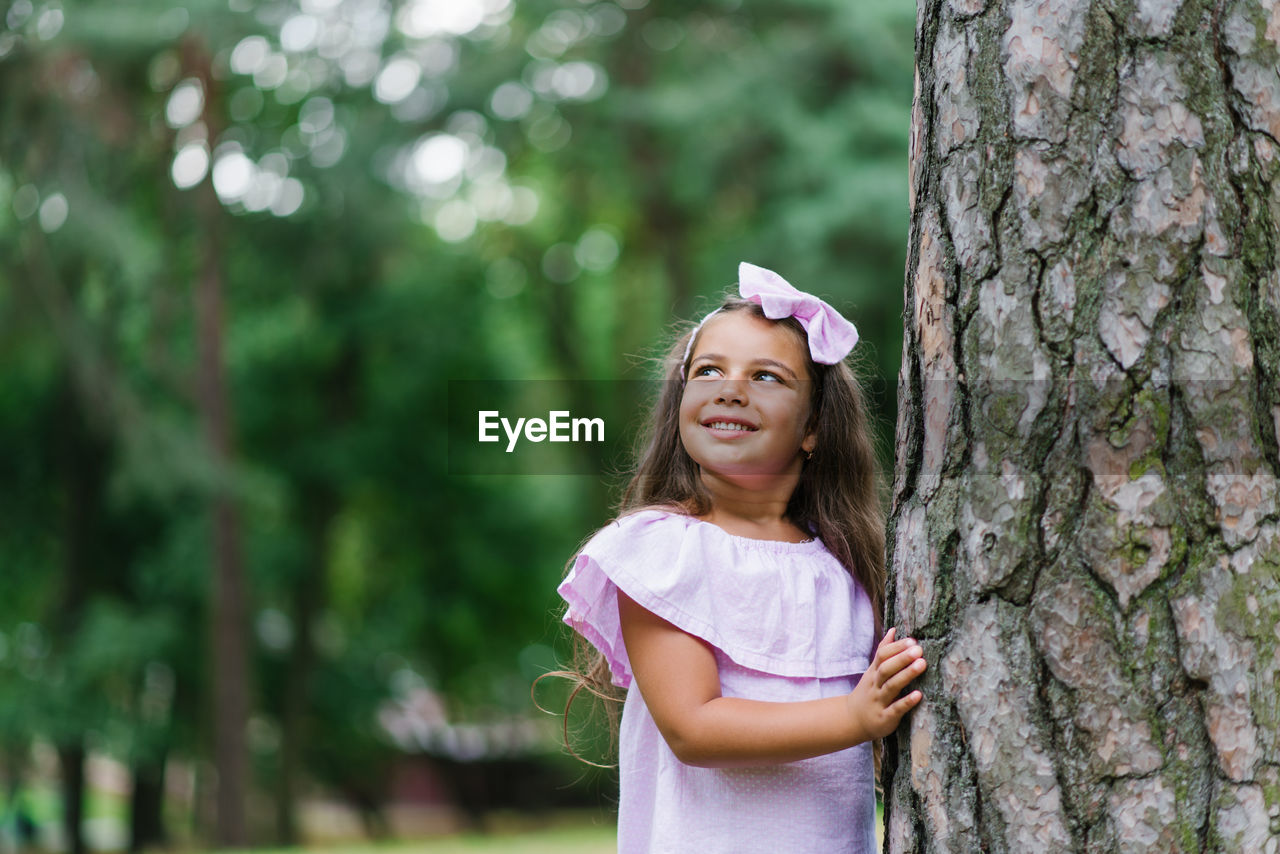 A beautiful girl in a pink dress stands near a tree in the forest in summer, smiling and dreaming