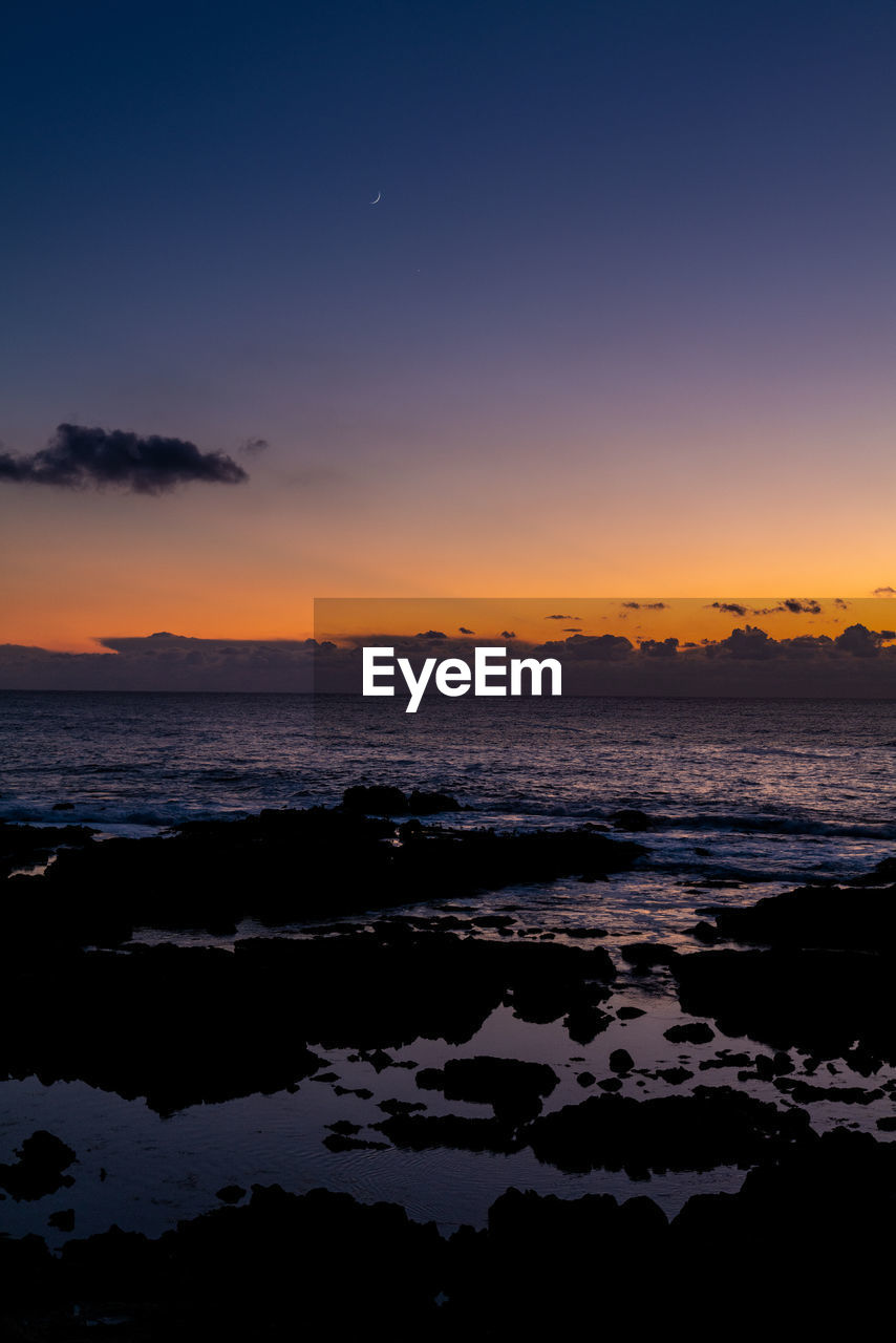 Crescent moon in colorful sunset sky above tide pools at edge of ocean