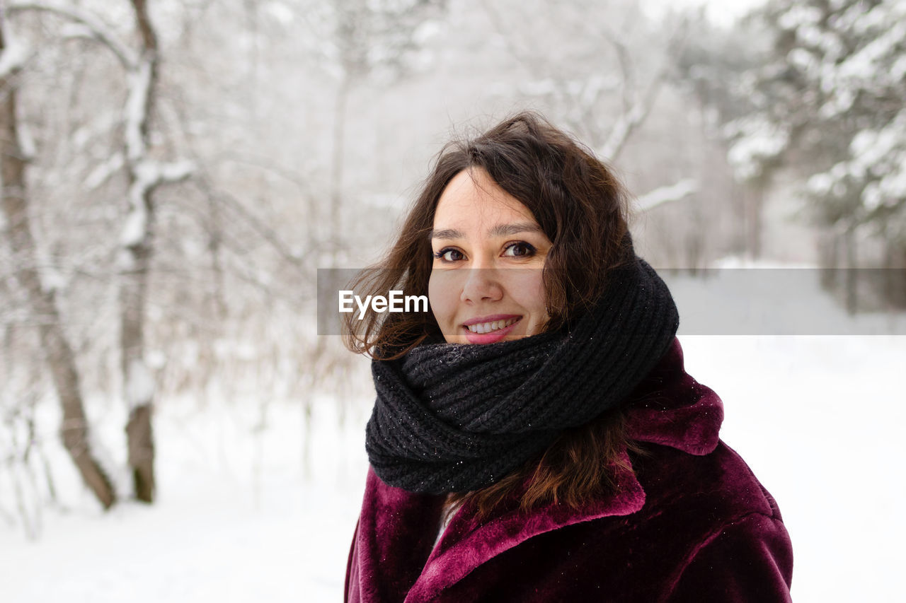 Smiling woman with long brown hair in a coat from faux fur on a background of winter forest.