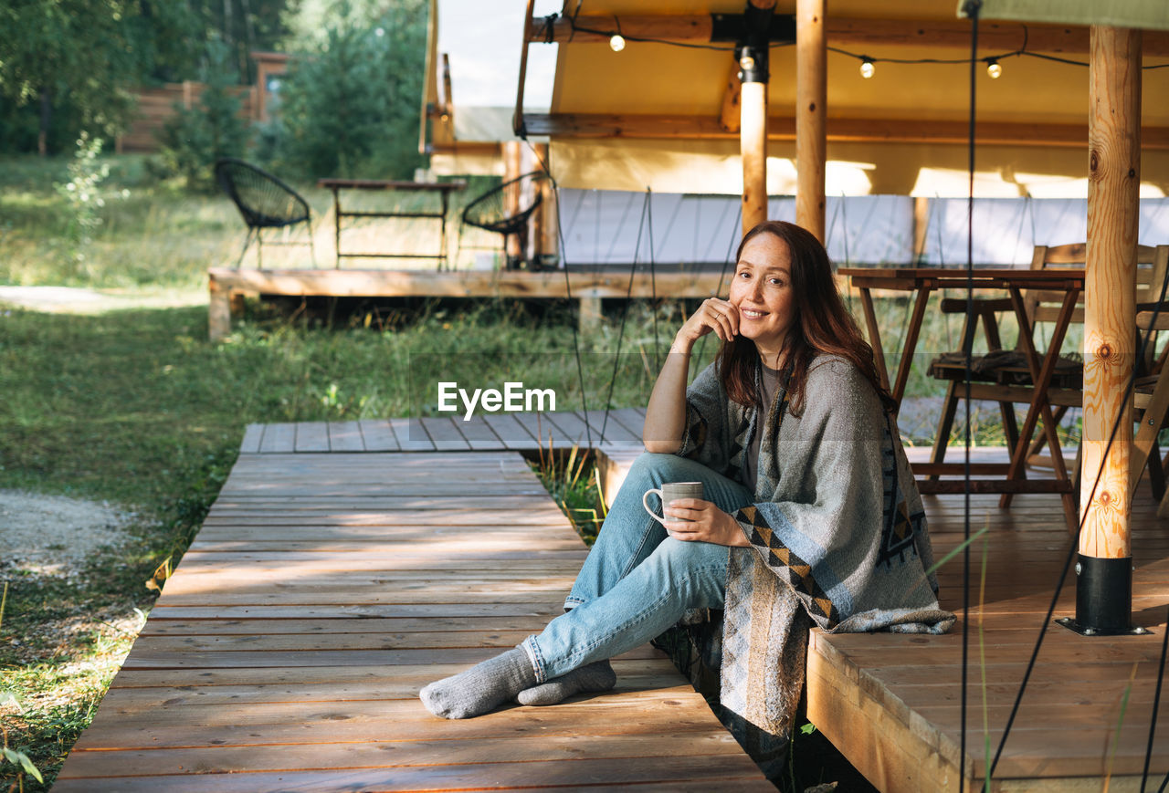 Young brunette woman in poncho drinking tea and relaxing in glamping in nature