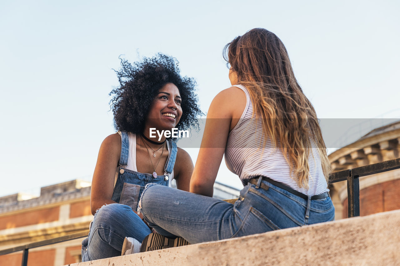 Female friends sitting on retaining wall against clear sky