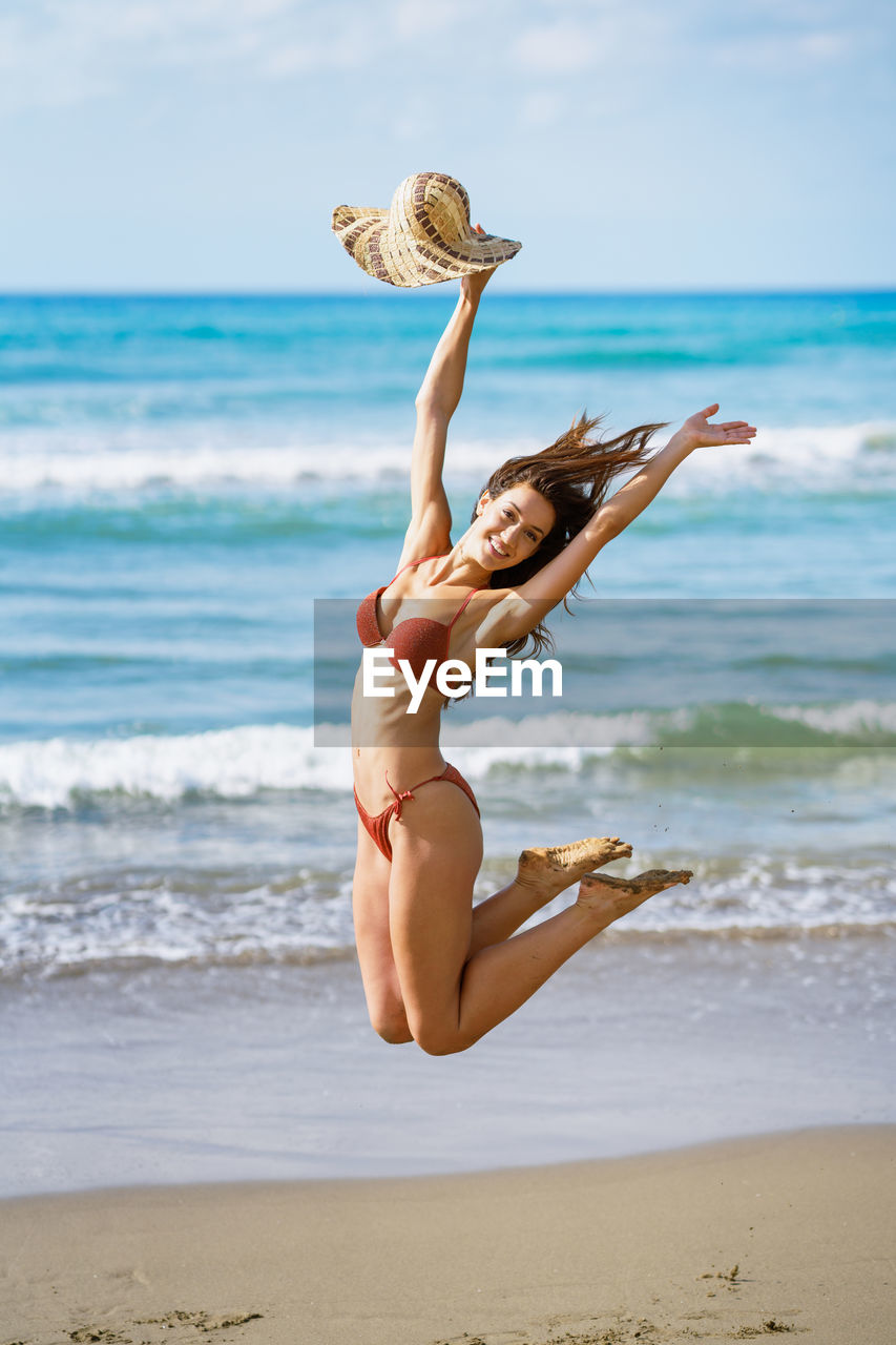 Full length of woman holding hat jumping on beach against sea