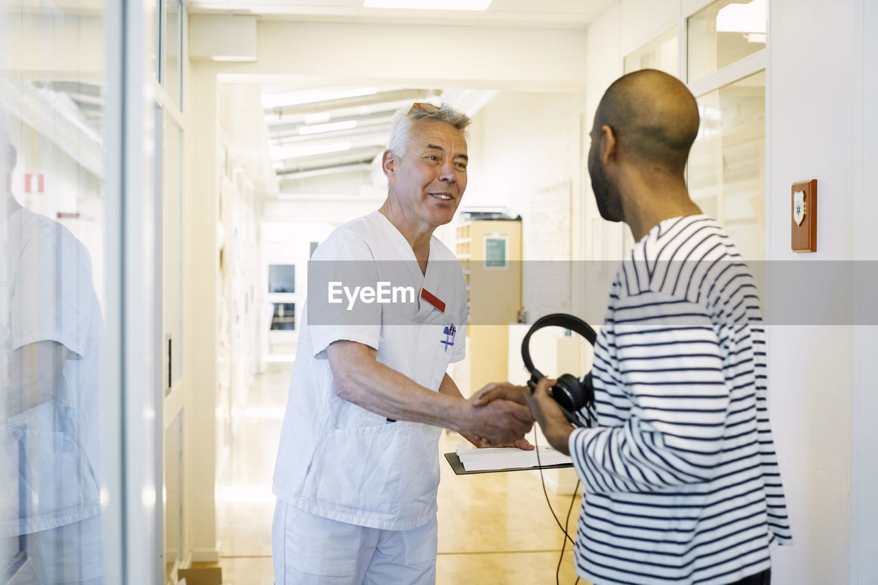 Senior doctor with clipboard greeting young man at hospital corridor