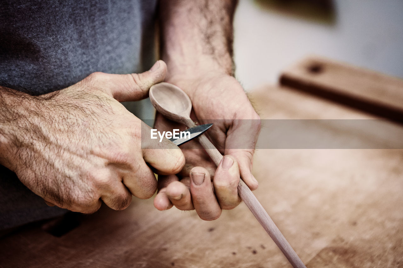 Midsection of carpenter carving wooden spoon with knife in workshop