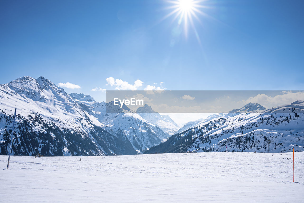 Scenic view of shining sun through blue sky over snow covered mountains