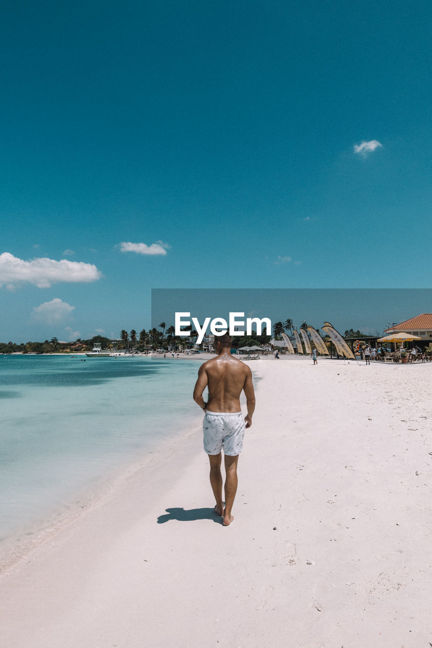 Rear view of shirtless man walking on shore at beach against blue sky