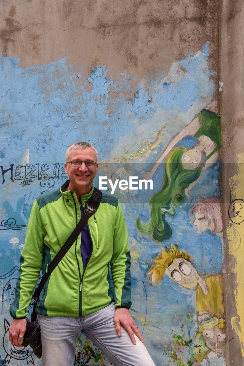 PORTRAIT OF SMILING YOUNG MAN STANDING AGAINST WALL