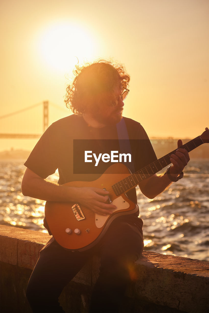 side view of man playing guitar while standing at sunset