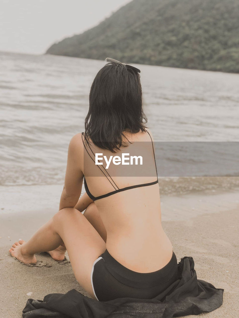 REAR VIEW OF WOMAN SITTING ON SHORE AT BEACH