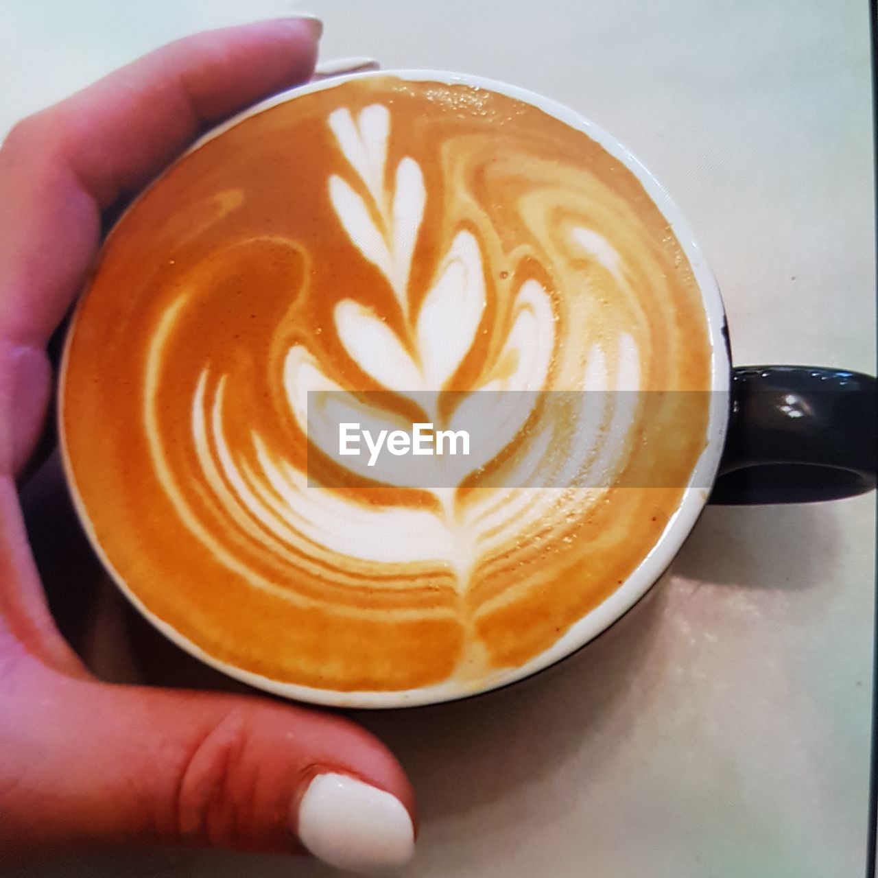 CLOSE-UP OF HAND HOLDING CAPPUCCINO WITH COFFEE CUP ON TABLE