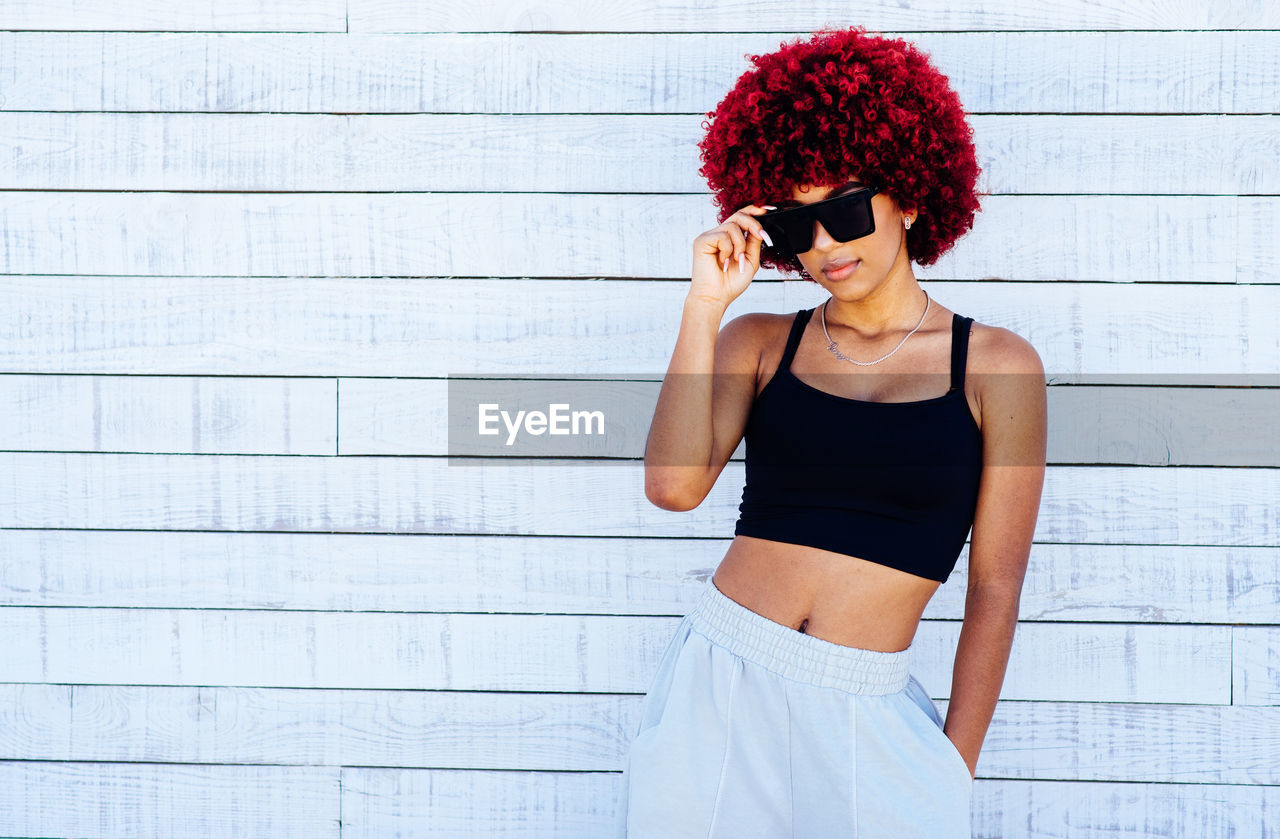 Woman with red afro hair and sunglasses standing on a white wall.
