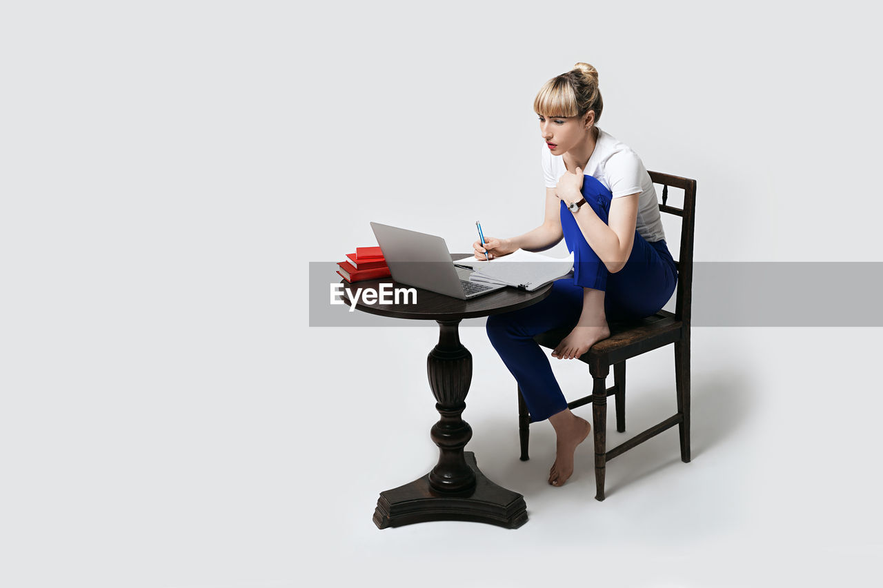 portrait of smiling young woman using mobile phone while sitting on chair against white background