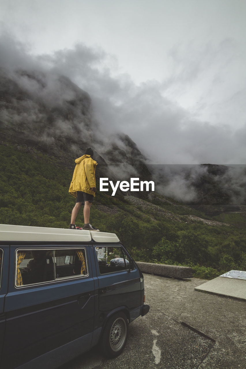 Rear view of man standing by mountain on vehicle during foggy weather