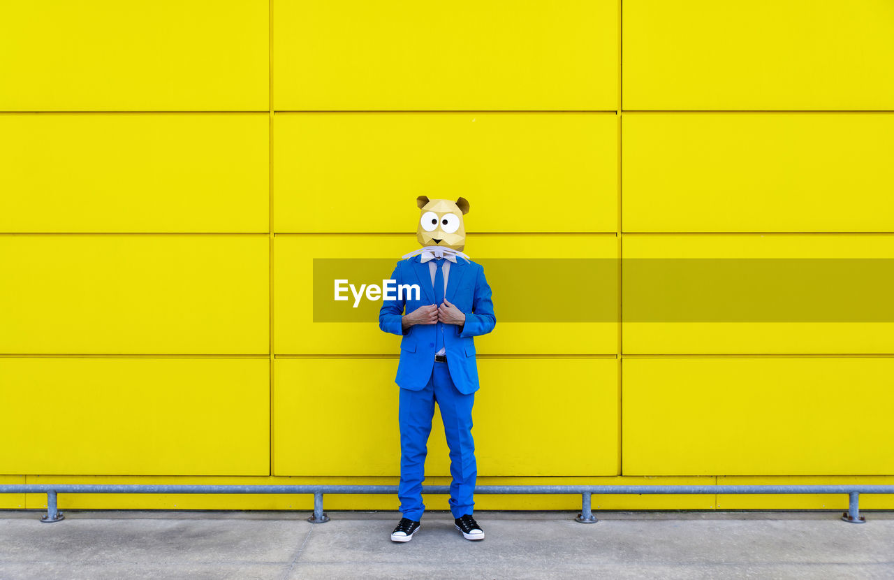 Man wearing vibrant blue suit and rodent mask standing in front of yellow wall
