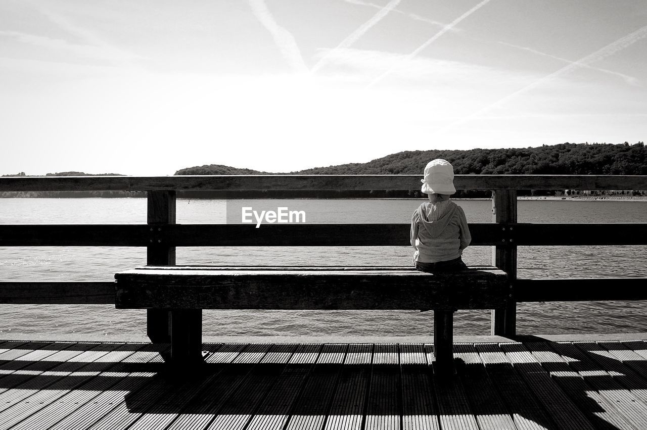 Girl sitting on bench at shore against sky