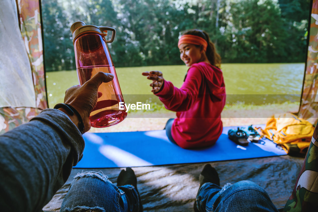 Midsection of man holding bottle with woman sitting in background