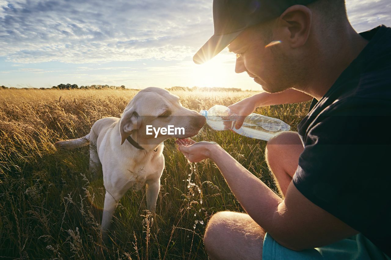 Man feeding water to dog on field against sky