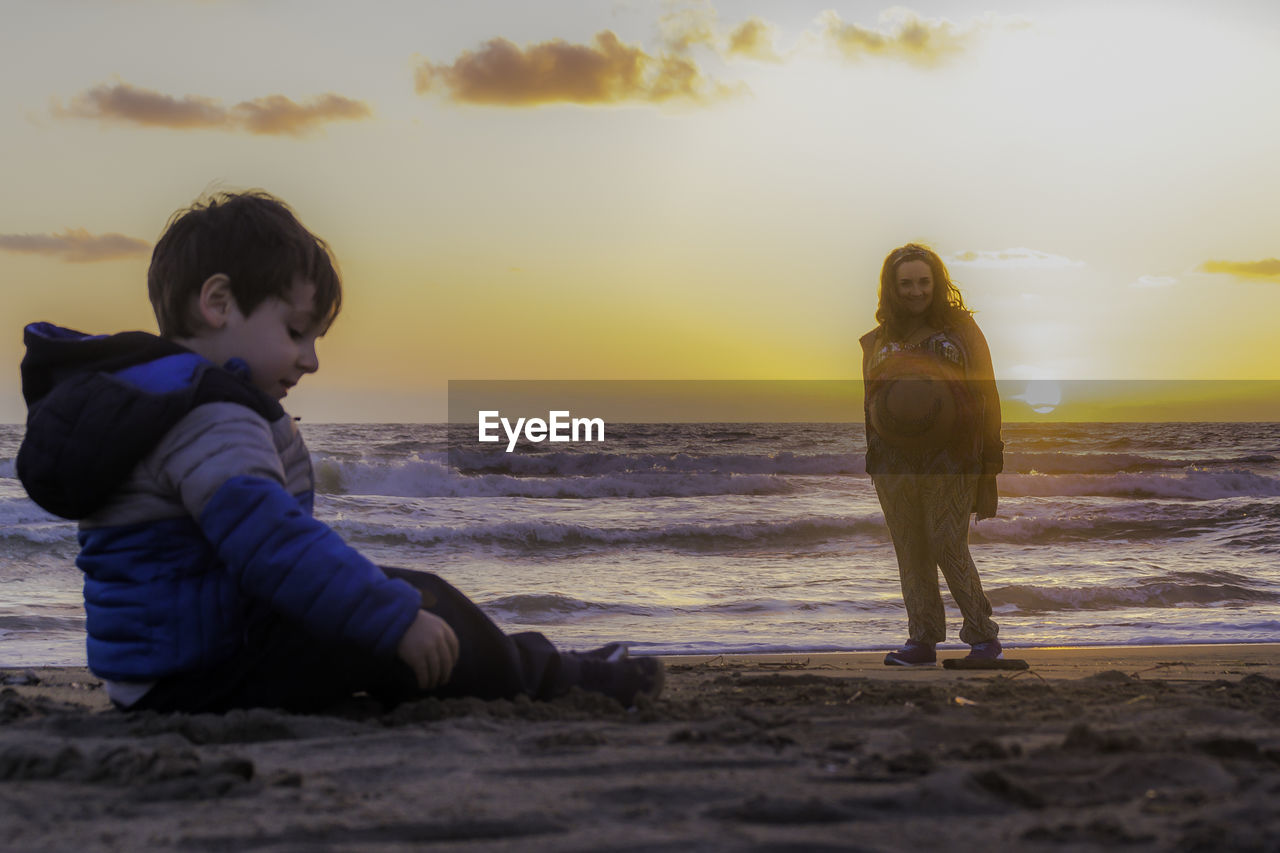 Full length of woman and boy on beach against sky during sunset