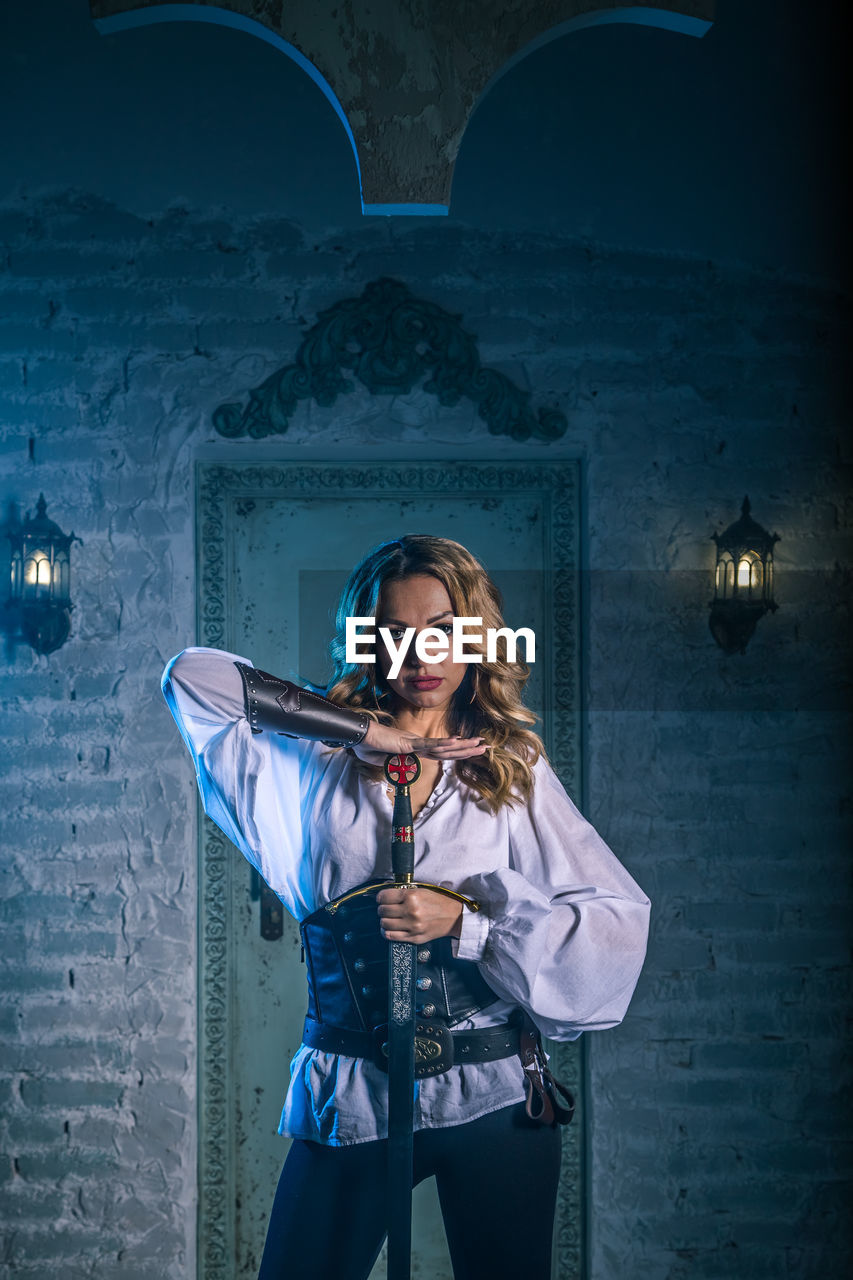 Portrait of woman in costume holding sword standing against door of abandoned building at night