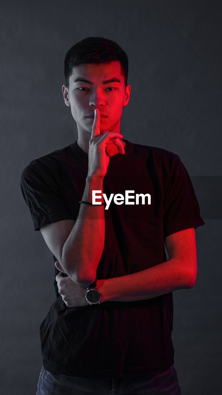 Portrait of young man with finger on lips standing against black background