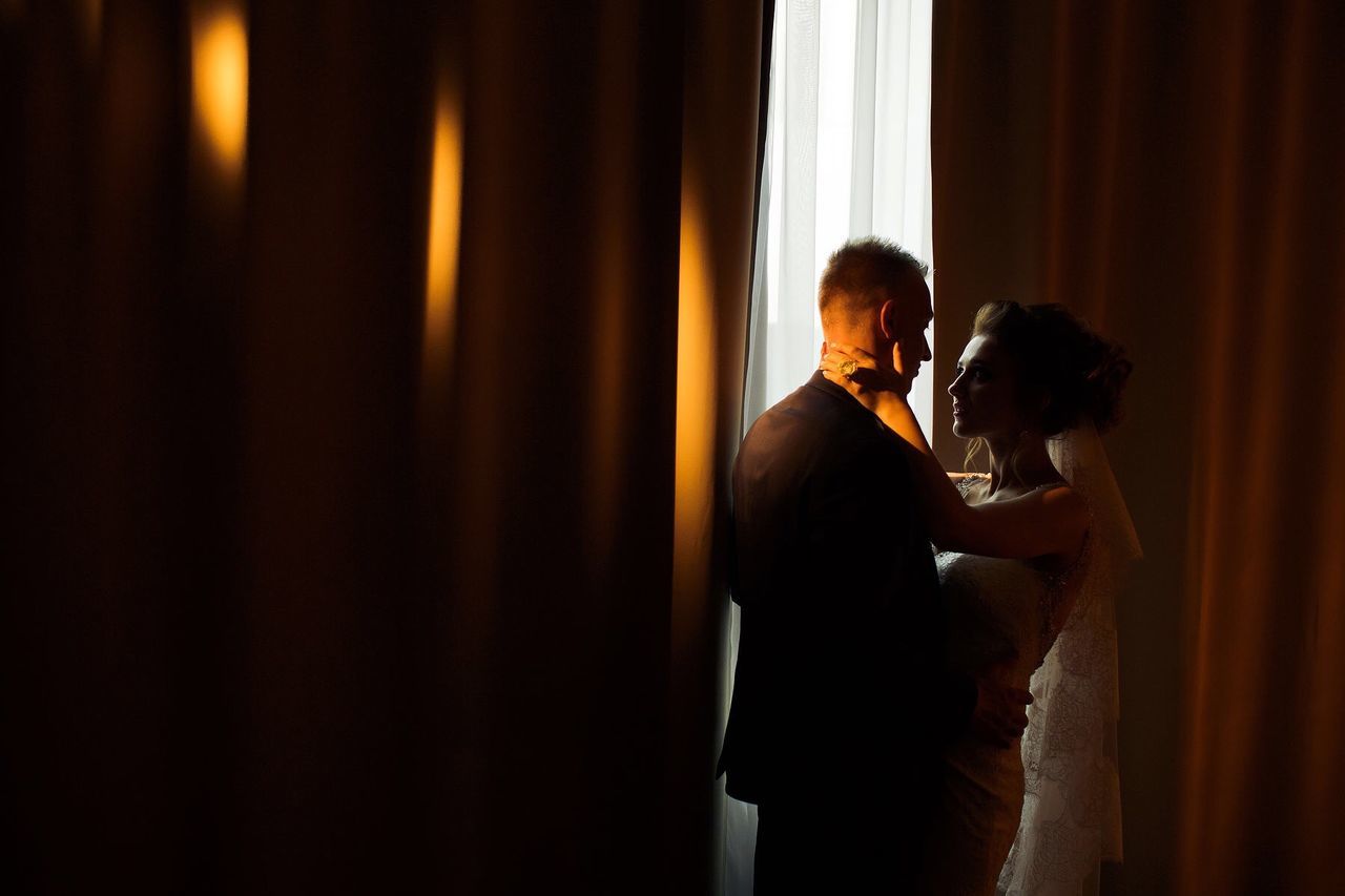 Romantic newlywed couple embracing in room against curtains