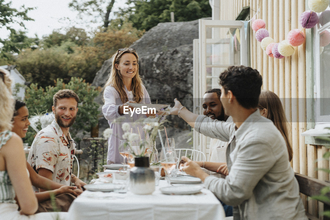 Smiling woman passing plate to male friend sitting at table during dinner party outside cafe