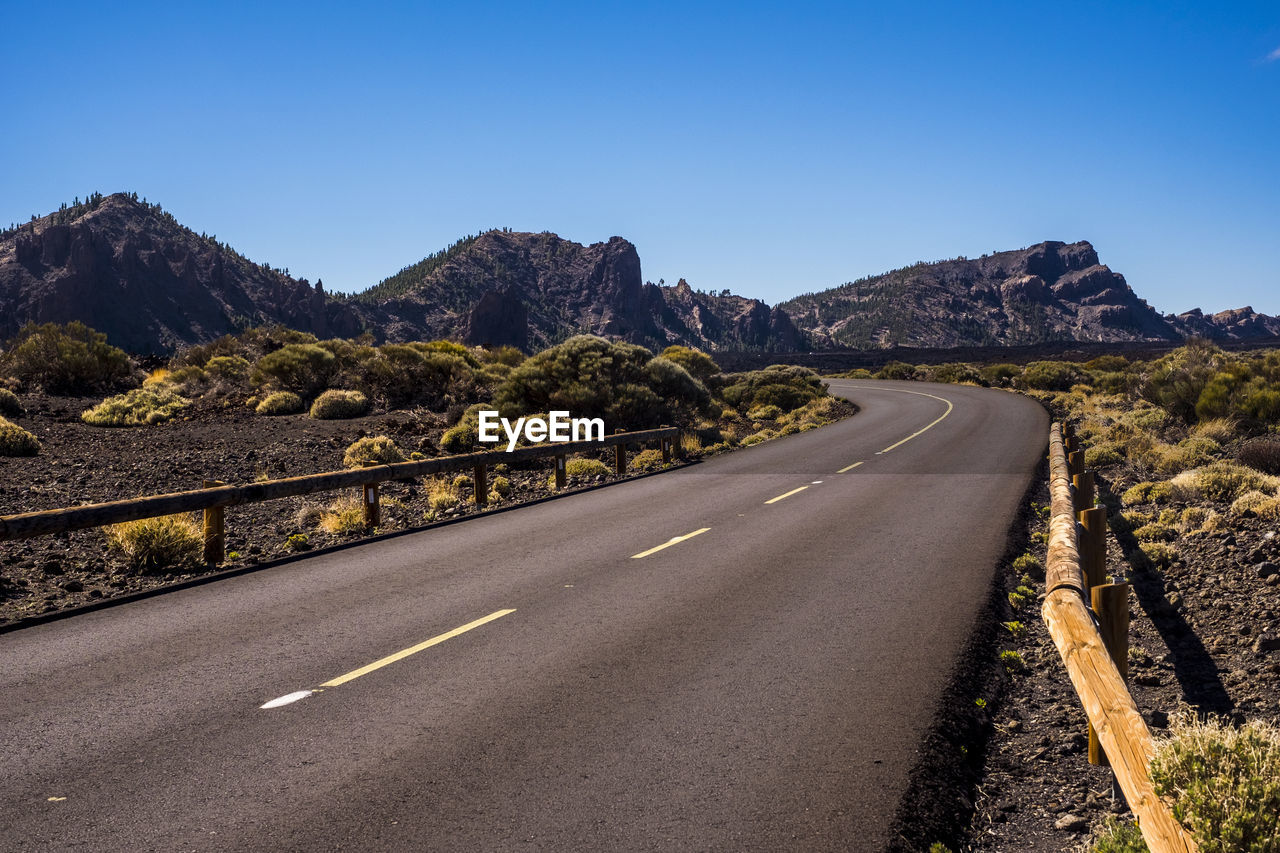 Road leading towards mountains against clear blue sky