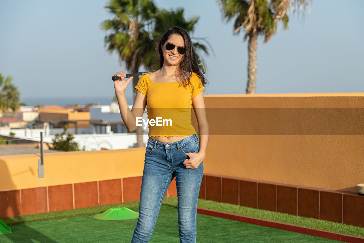 one person, fashion, sunglasses, adult, young adult, yellow, palm tree, glasses, casual clothing, women, standing, smiling, hairstyle, portrait, architecture, tropical climate, happiness, nature, leisure activity, lifestyles, plant, tree, emotion, long hair, day, front view, full length, sky, brown hair, sunlight, outdoors, jeans, clothing, summer, cheerful, looking at camera, city, cool attitude, enjoyment, built structure, fun, copy space, looking, sunny, relaxation, building exterior, holding, person