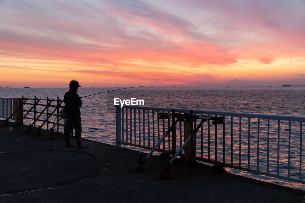 sunset, sky, sea, water, horizon, evening, ocean, pier, dusk, railing, nature, beauty in nature, beach, scenics - nature, cloud, full length, horizon over water, coast, land, silhouette, tranquility, vacation, architecture, trip, adult, one person, holiday, shore, travel, tranquil scene, orange color, travel destinations, standing, leisure activity, lifestyles, idyllic, outdoors, twilight, men, sun, tourism, relaxation, dramatic sky, city, sunlight, transportation, walking, women, activity, built structure