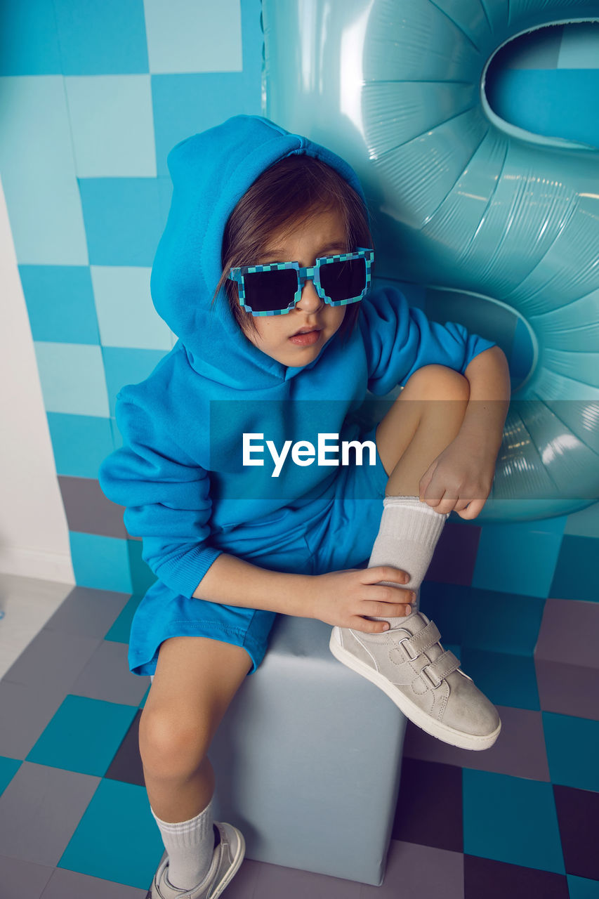 Boy child in blue clothes and a cap is sitting on a gray square celebrating his birthday.