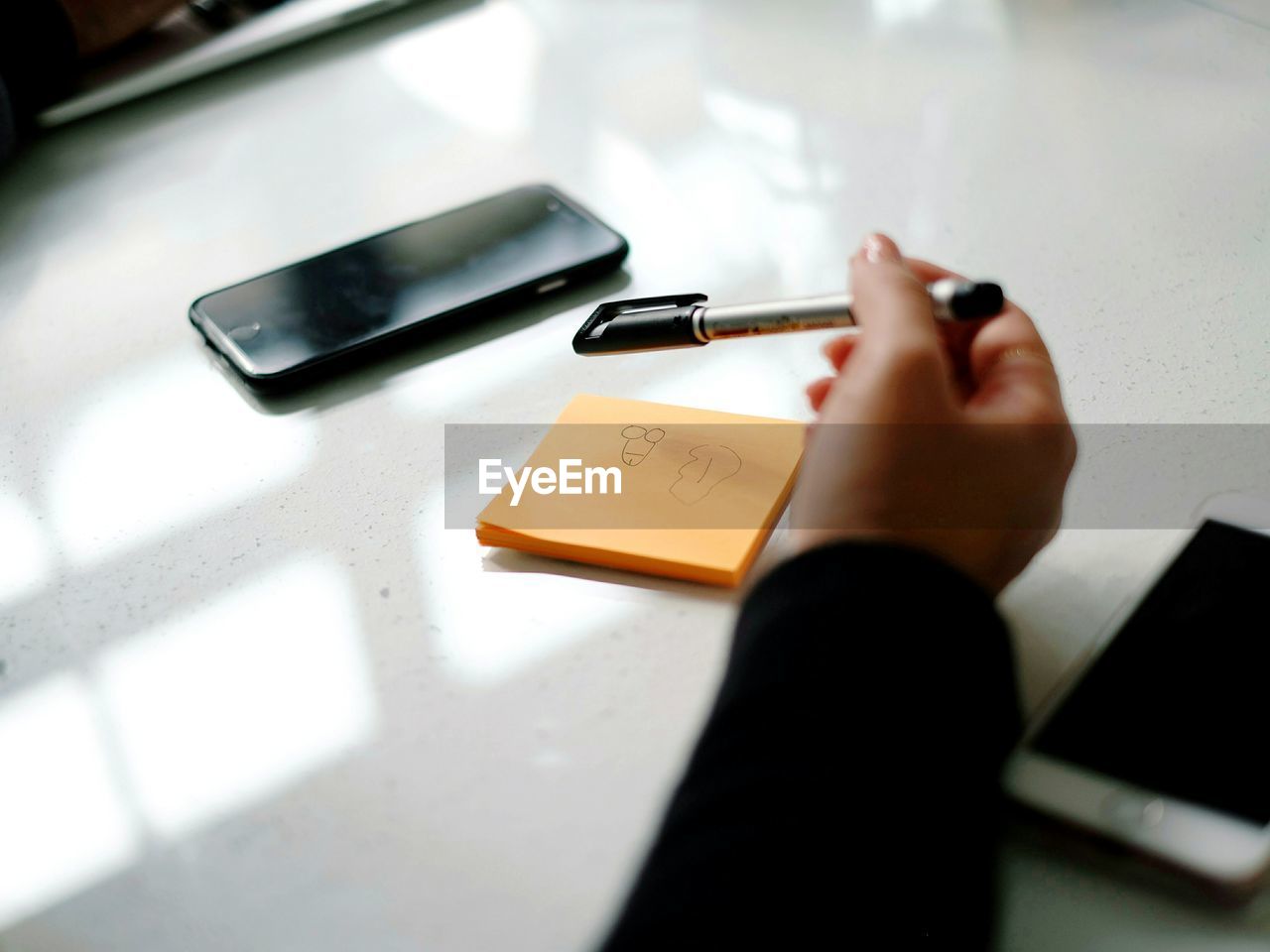 Cropped hand of businesswoman holding pen over sticky notes by mobile phones on table