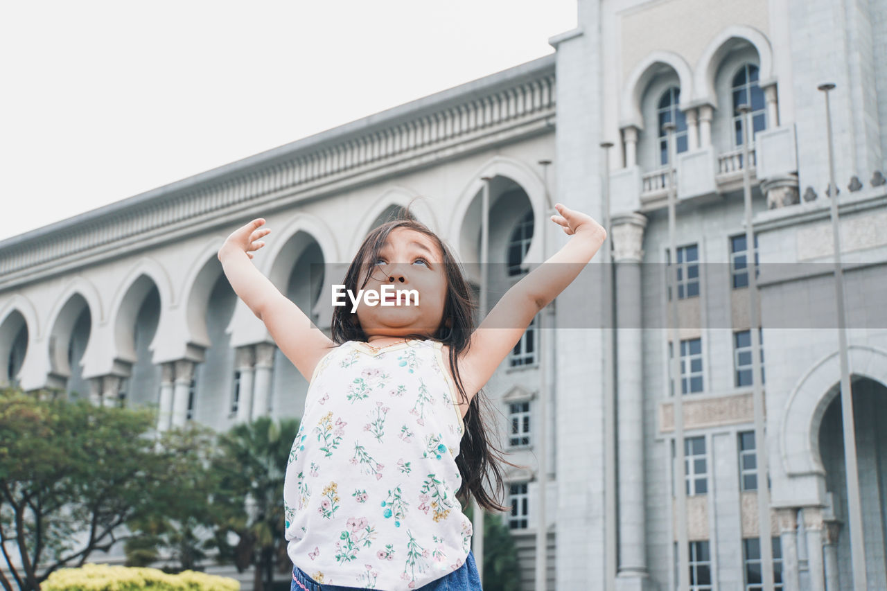 Cute girl standing with arms raised standing against building in city