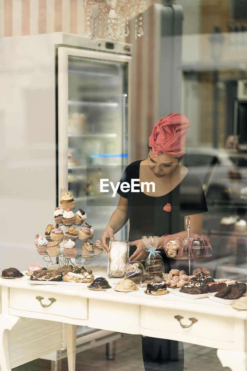 Hispanic woman in headscarf arranging desserts on table near window while working in bakery in daytime