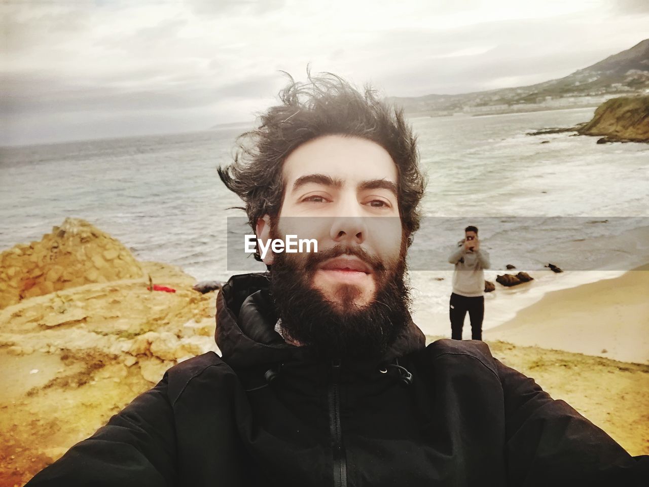 Portrait of young man standing at beach against cloudy sky