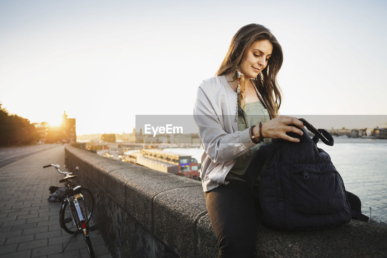 Female tourist searching in backpack while sitting on retaining wall of bridge against clear sky