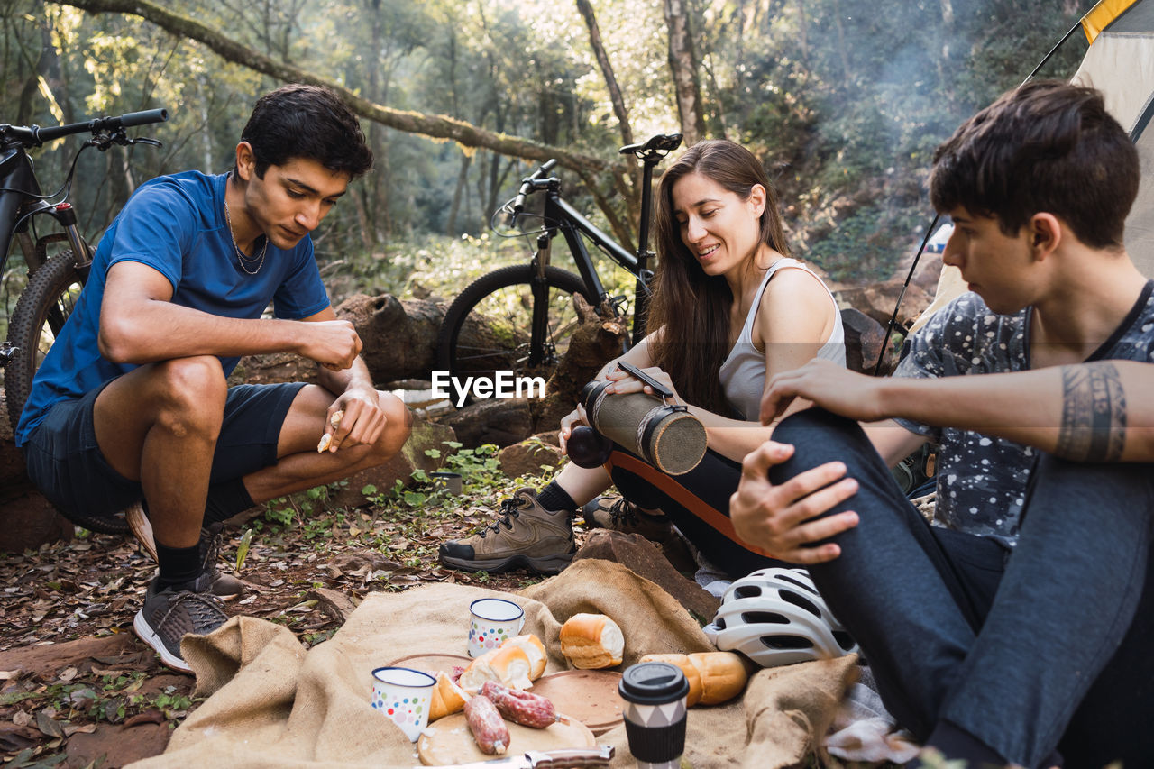 Diverse cyclists sitting on ground near tent and having picnic while hiking together in forest