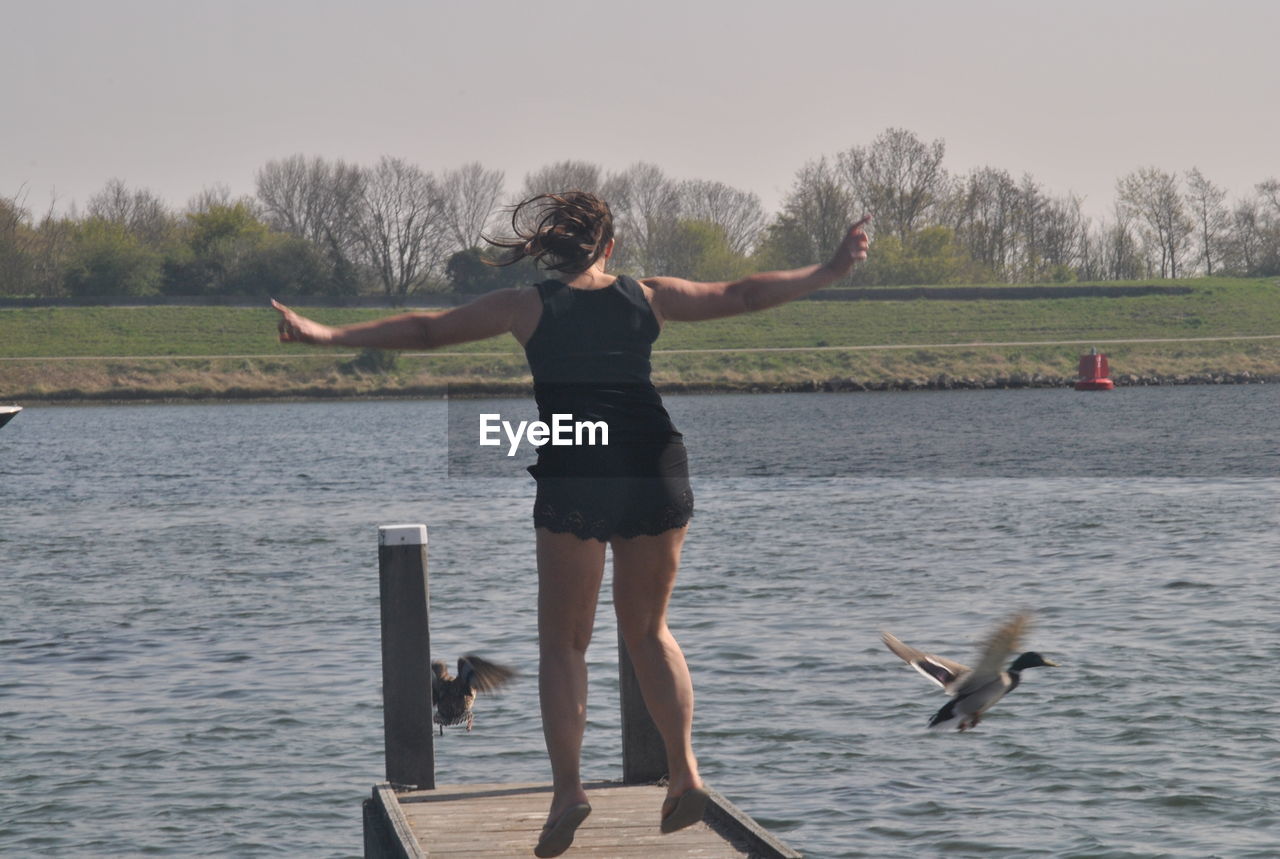 Rear view of young woman jumping on pier over lake against sky