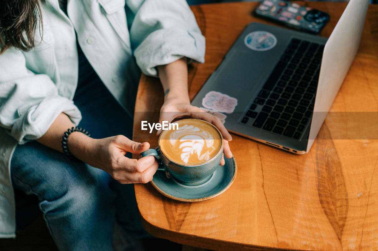 Female hands hold a large mug with cappuccino at a table in a cafe next to a laptop.