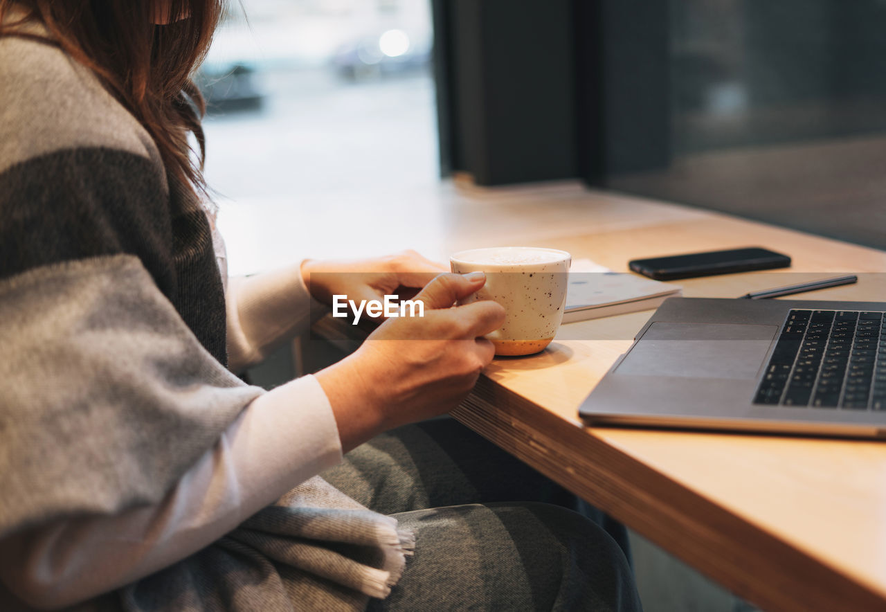 Crop photo of brunette woman in warm scarf working with laptop on table at cafe in the cold season