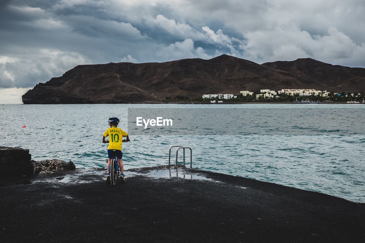 Rear view of boy on bicycle by sea against cloudy sky