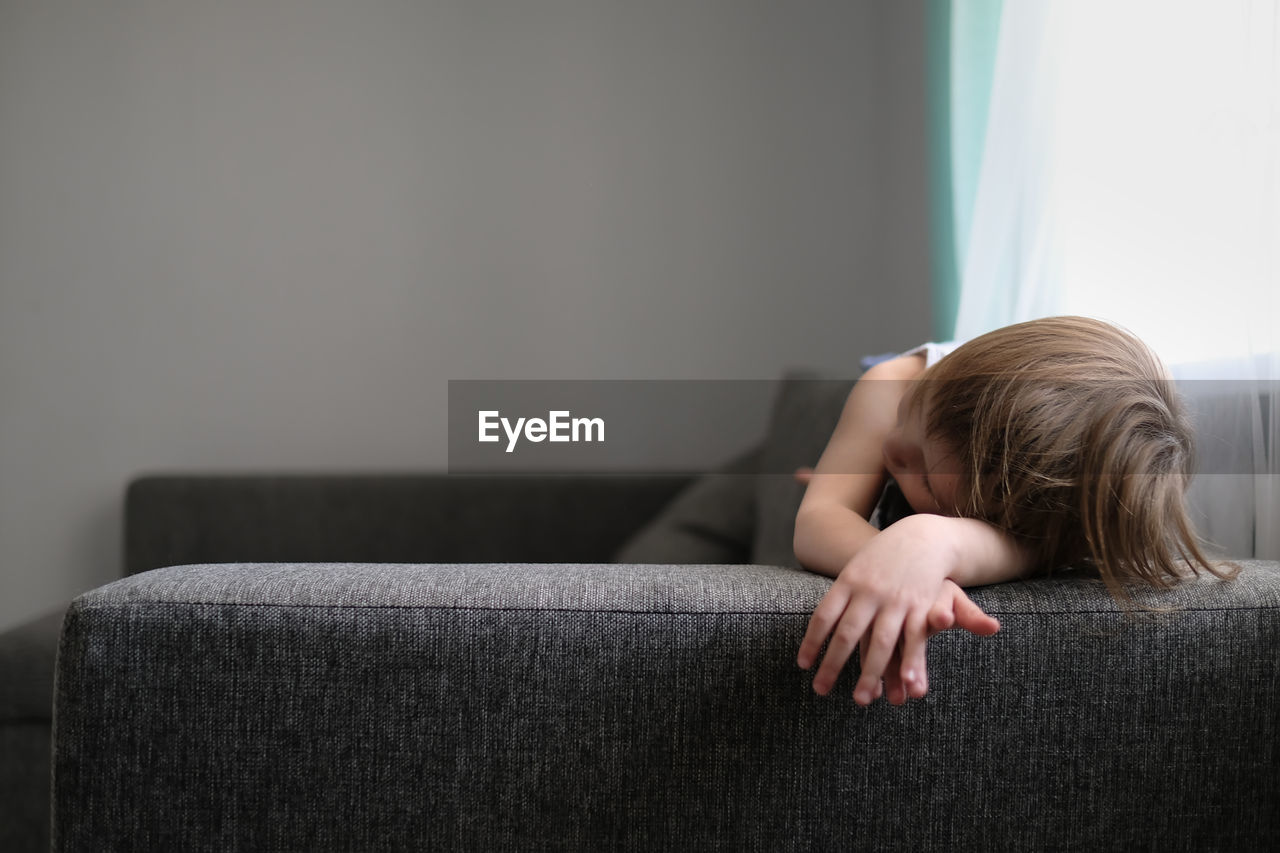 European toddler child sits and gets bored on gray sofa in bright living room, minimalist interior