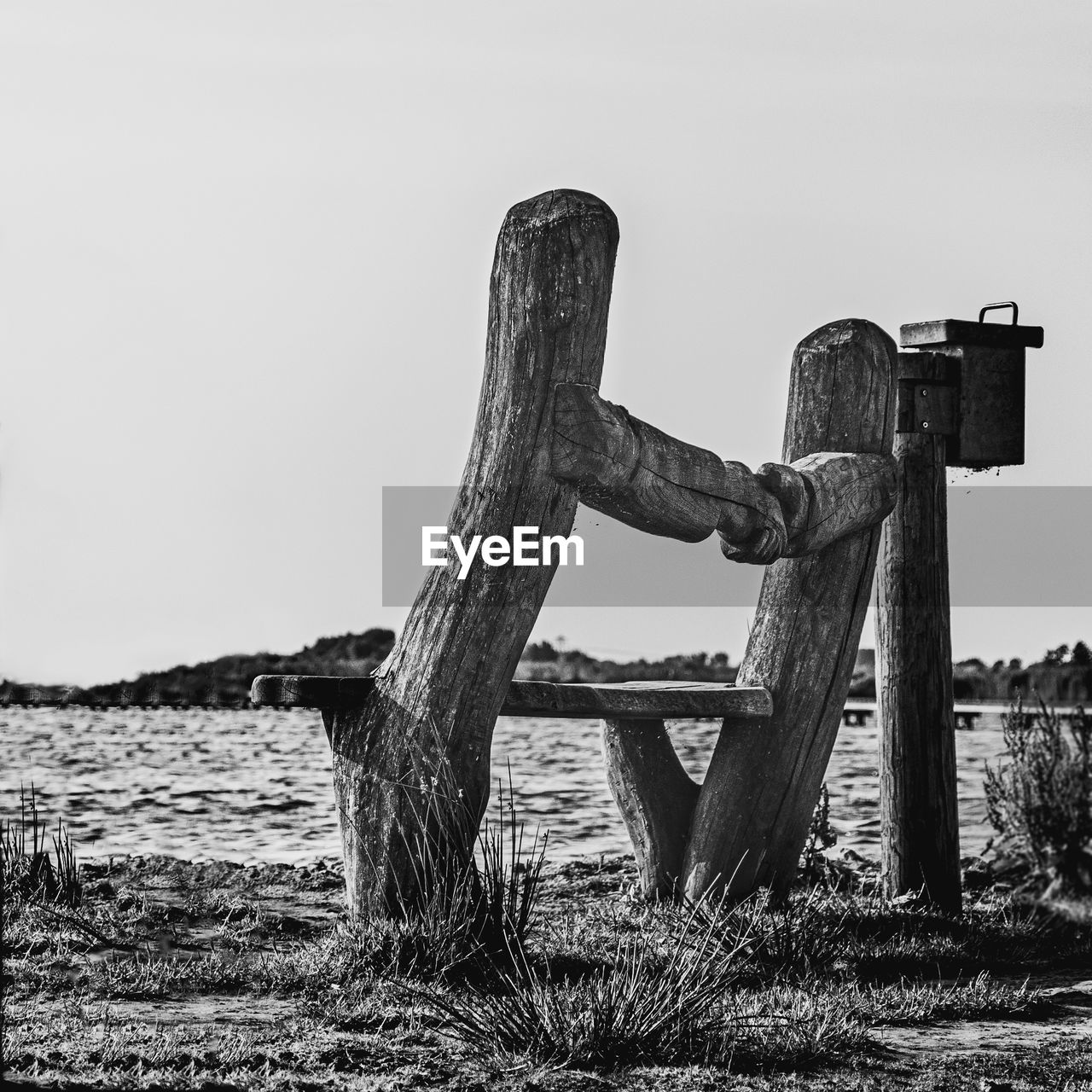 Bench by the lake with attached postbox surrounded by low grass in black and white 