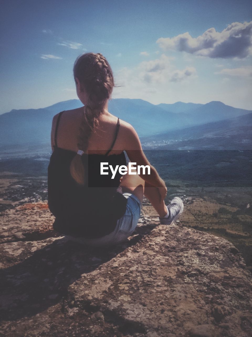 Rear view of woman sitting on cliff against sky during sunny day