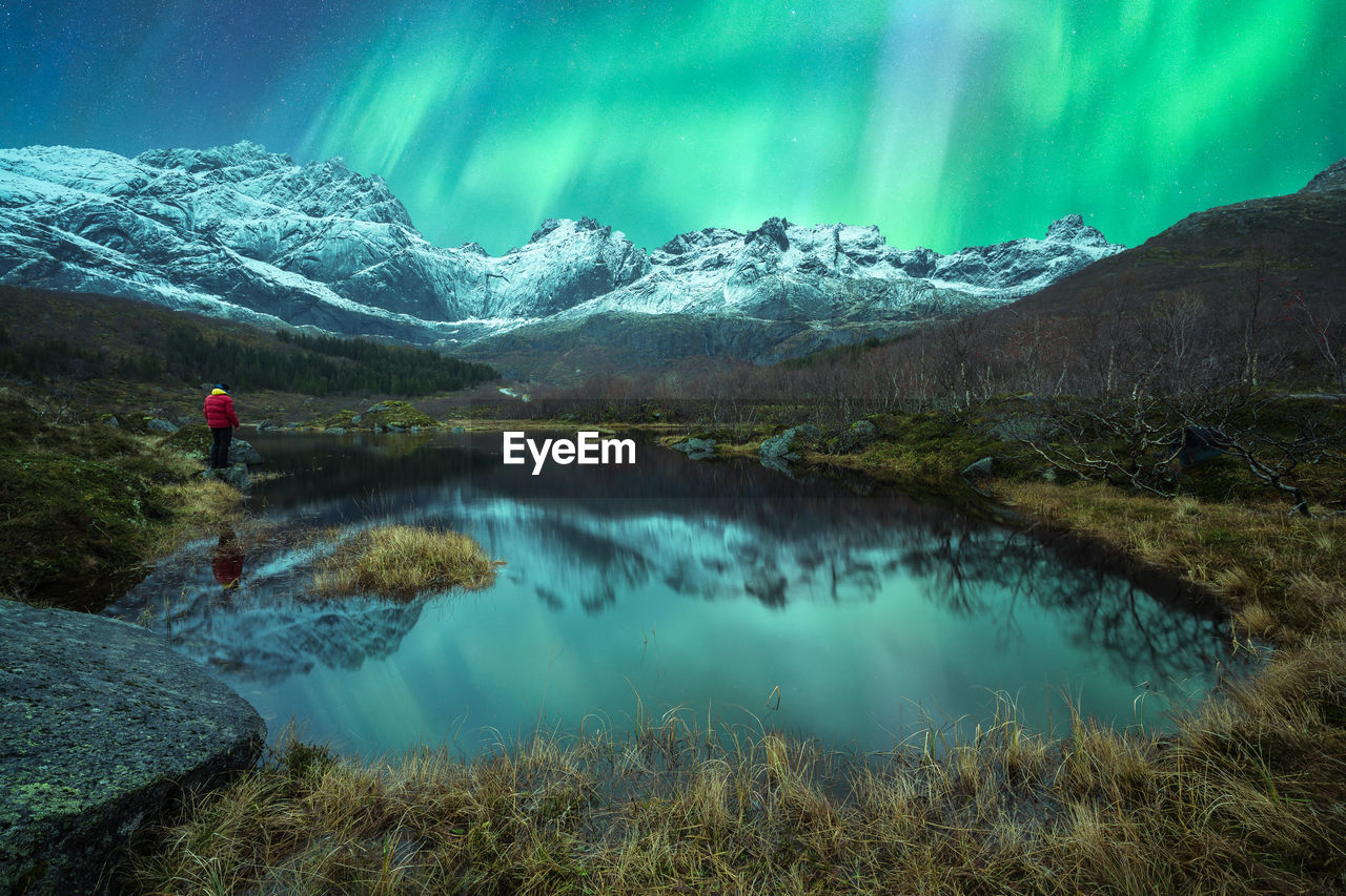 Unrecognizable traveler standing near pond in rocky snowy mountains and admiring view of green aurora borealis illuminating dark night sky in lofoten islands