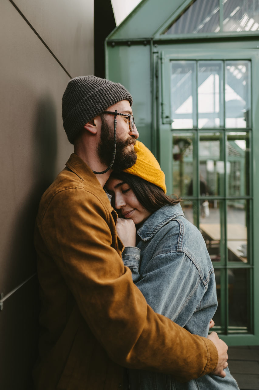 Side view of young stylish couple hugging gently while standing on wooden terrace near house