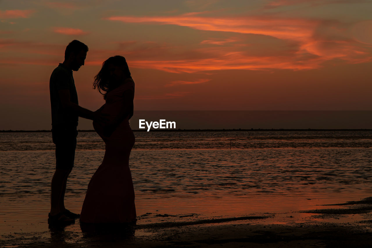 SILHOUETTE FRIENDS STANDING AT BEACH AGAINST SKY DURING SUNSET