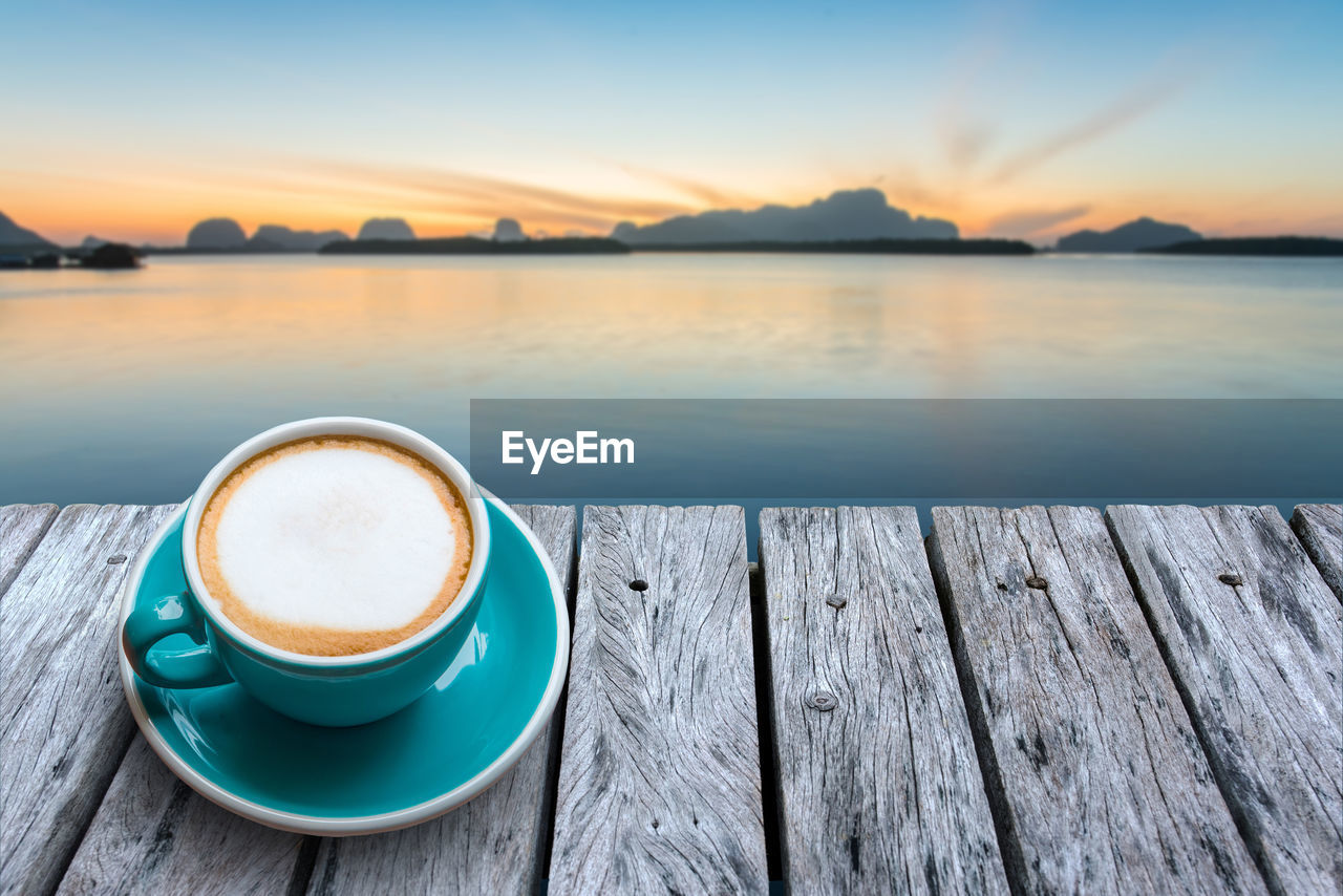 Fresh coffee in cup on old wooden table and calm sea and beautiful sunrise.