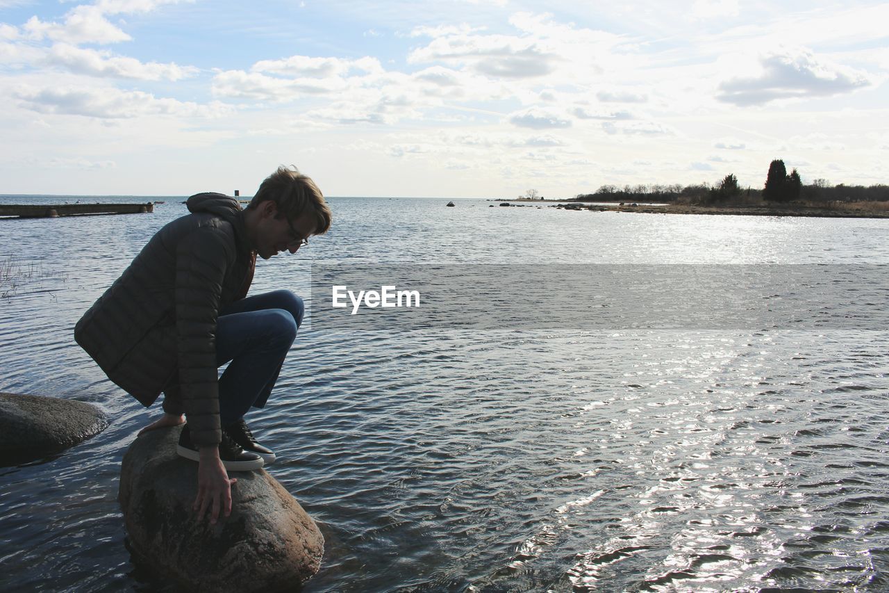 Side view of man crouching on rock in lake against sky